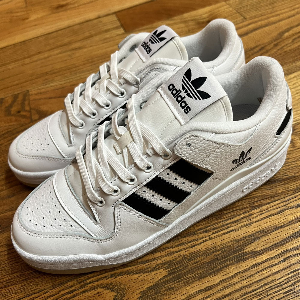 A pair of Forum 84 Low Adidas Skateboarding Cloud White/Core Black leather shoes with black stripes and logos, perforated toes, white laces, and branding on the tongue, side, and heel rest on a wooden floor.