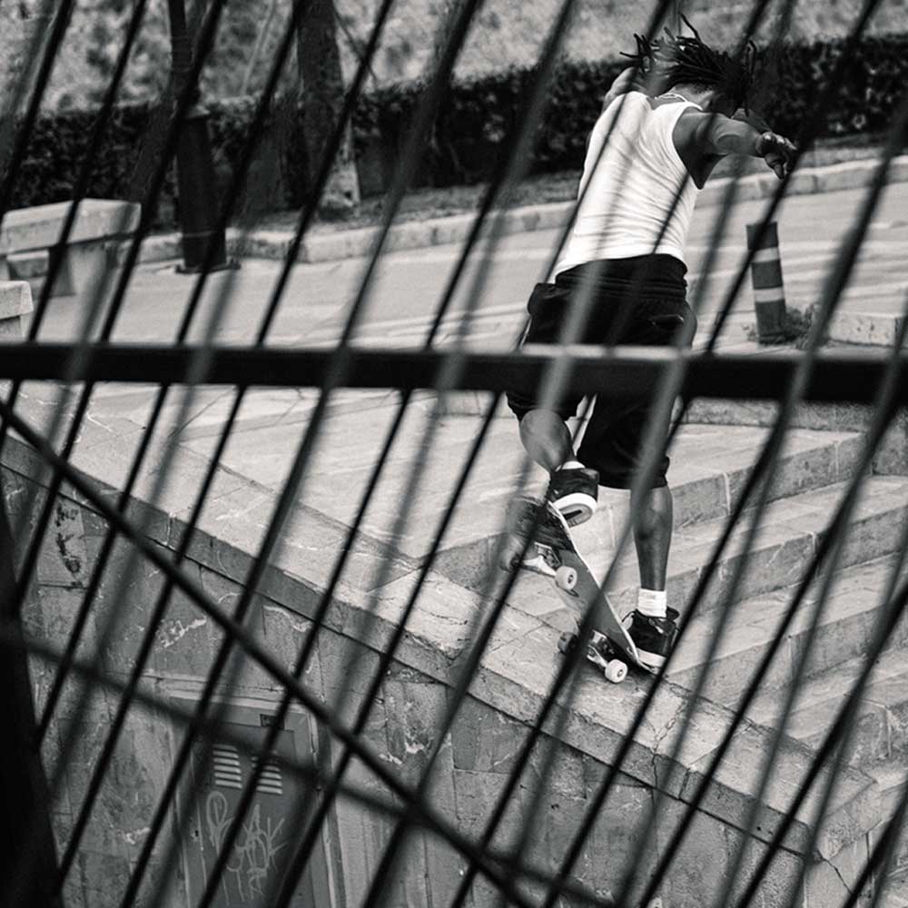 A person in a white shirt and shorts skateboards down concrete steps in Superskate x Kader Shoes JQ4413 Core Black/Off White Leather Suede Adidas Skateboarding Shoes—dynamic black and white photo seen through blurred metal bars.
