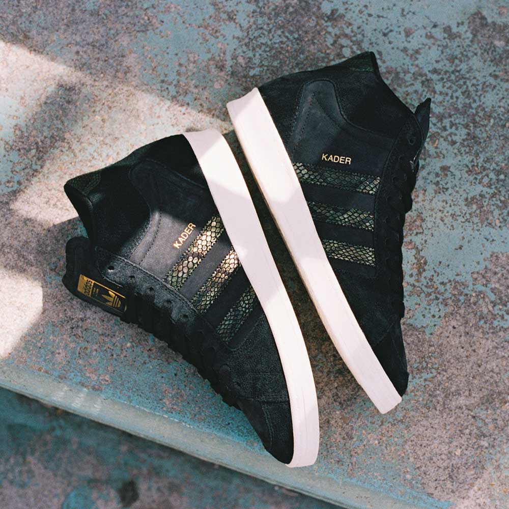 A pair of adidas Superskate x Kader Shoes (JQ4413) in core black with off white soles, leather and suede upper, "KADER" on the side, placed side by side on a textured concrete surface.