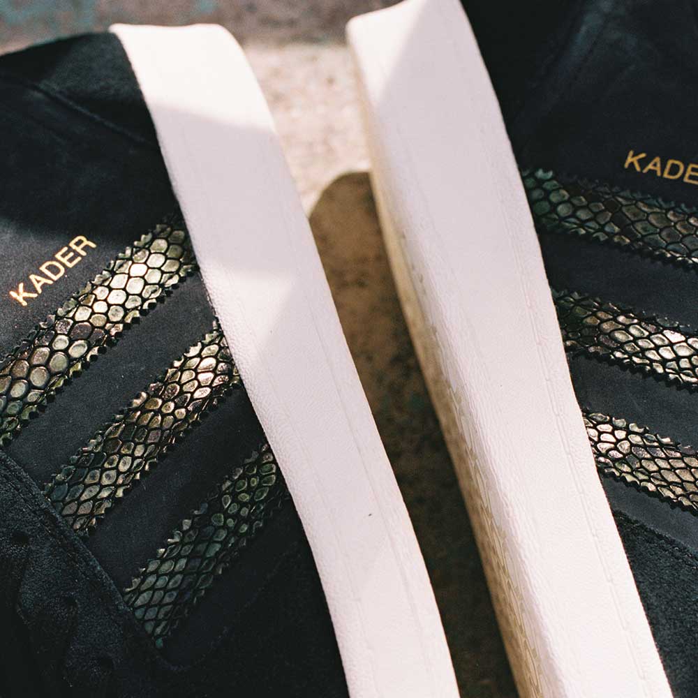 Close-up of two adidas Superskate x Kader Shoes (JQ4413) in core black with off-white soles, gold KADER text, a suede and leather upper, and reptile-scale-patterned stripes on the sides.