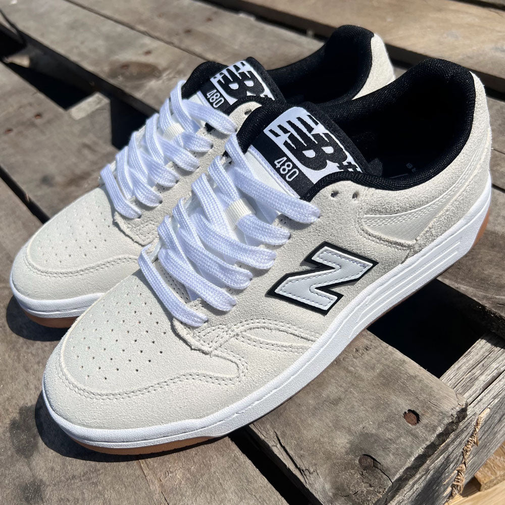 A pair of 480 SWG New Balance Numeric Shoes Suede White Black are side by side on a wooden pallet under sunlight.
