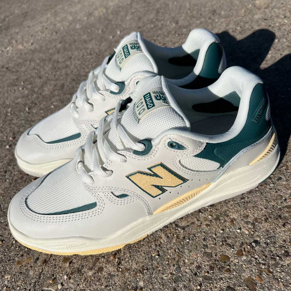The 1010 AL New Balance Numeric Suede Shoes in white, green, and yellow are shown from a side angle on a textured outdoor surface in bright sunlight.