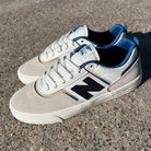 A pair of 306 TWC Jamie Foy New Balance Numeric Suede Shoes in white, blue, black, and brown sits on a textured concrete surface in sunlight.