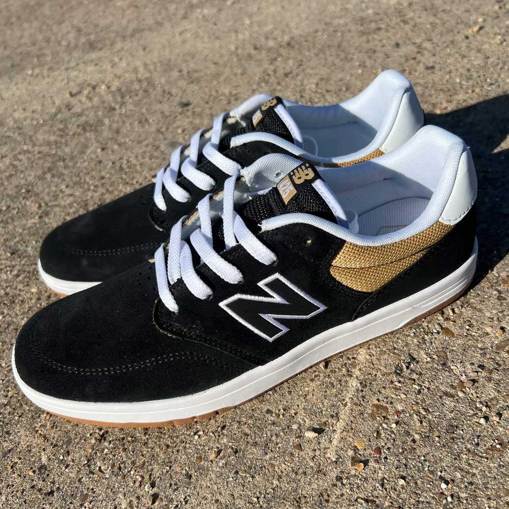 A pair of 425 BNT New Balance Numeric Suede Shoes in black with white soles, white laces, and gold accents, featuring C-CAP midsole cushioning, is shown on a rough concrete surface under bright sunlight.