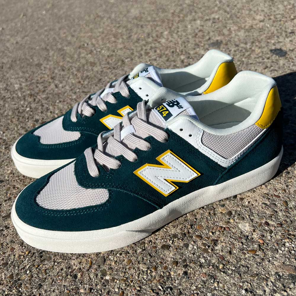 A pair of 574 VOK New Balance Numeric Gray White Suede Shoes featuring dark green suede, gray mesh panels, white soles, yellow accents, and a yellow-outlined N logo sits on a textured outdoor surface.