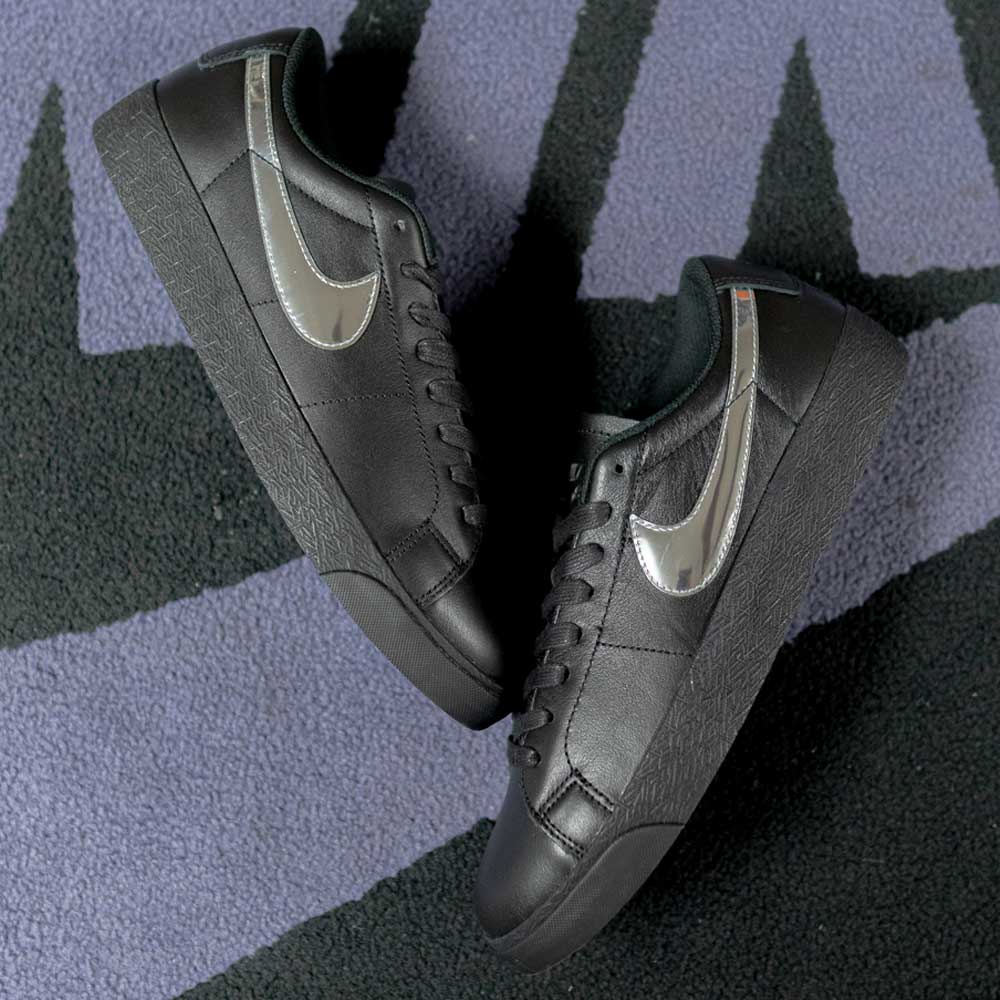 The HJ6703001 Black White Silver Blazer Low Dancer Nike SB leather skateboarding shoes feature metallic silver swooshes and are showcased on a surface with purple and black geometric textures—a stylish collab with Dancer.