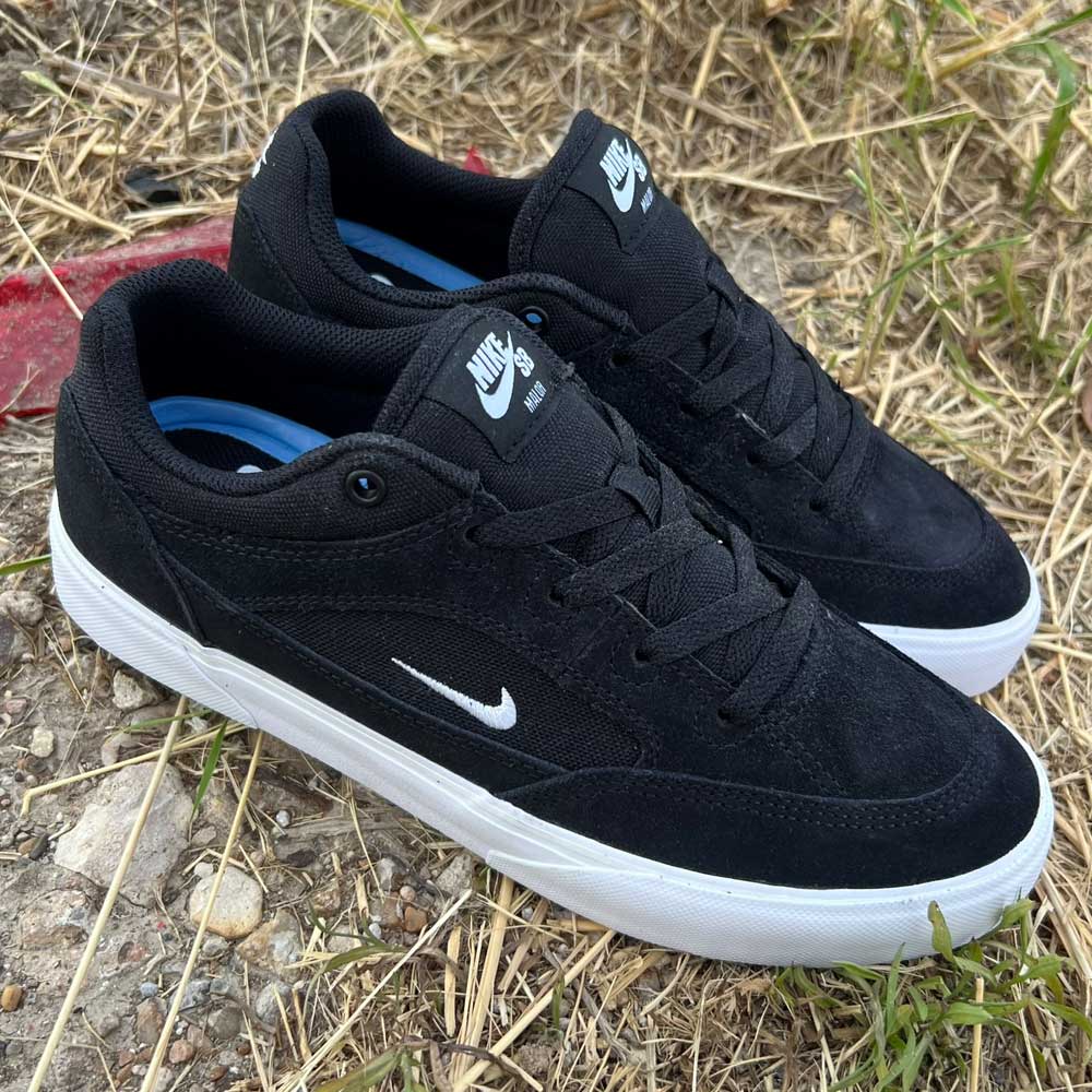 A pair of FV6064001 Black White Malor Nike SB suede skateboarding shoes with white soles and swoosh logos lies on dry grass and gravel outdoors.