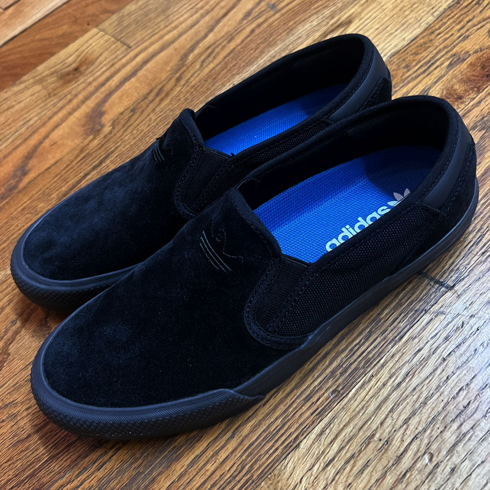 A pair of Shmoofoil Slip Adidas Skateboarding Core Black Carbon Suede Shoes with blue insoles and mesh sides rest on a wooden floor, displaying the Adidas logo inside.