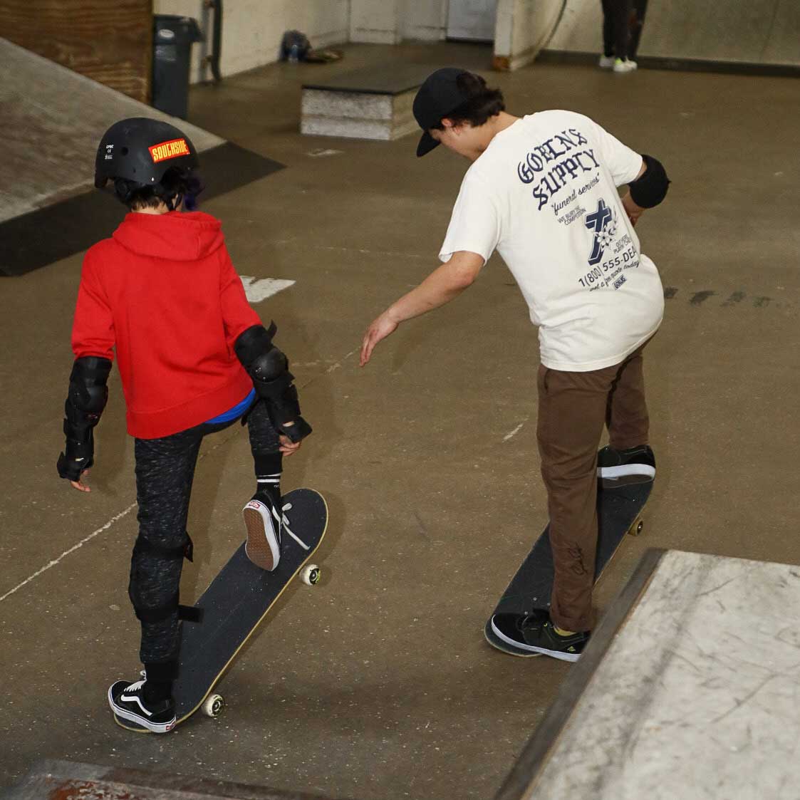 An adult teaches a child in a red hoodie and helmet skateboarding indoors at Southside Skatepark during the Instructional Spring Break Skateboarding Day Camp, held March 9th-11th, 2026. Both wear protective gear and balance on their boards together.