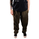 A person with tattoos on their left arm stands against a white background wearing a black t-shirt and loose-fitting Texas Icon Dark Green Southside 94 Pants, light-colored shoes, and their hands relaxed by their sides.