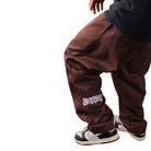 A person wearing Texas Icon Brown Southside 94 Pants featuring loose fit and white graffiti lettering on one leg, paired with a black shirt and black and white sneakers. Only the lower body is visible.