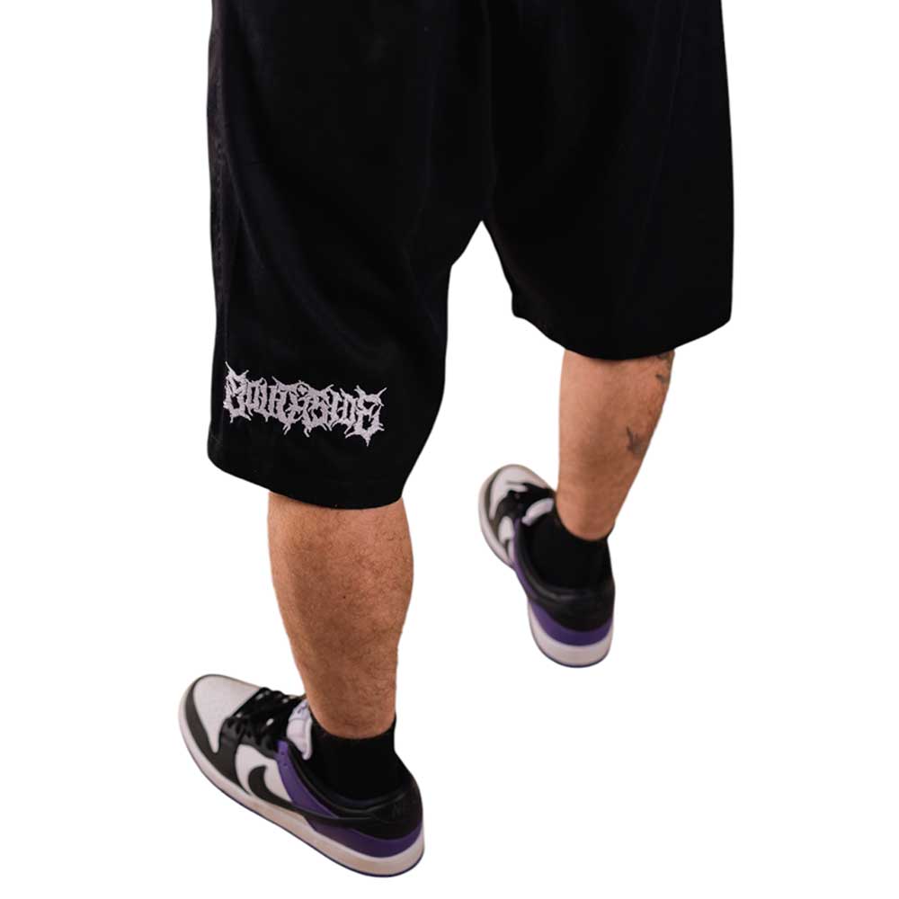 Someone wearing Texas Icon Black Southside 94 Shorts with bold white leg graphics and black-and-white sneakers with purple accents stands against a white background. Only lower legs and feet are shown, embodying classic skateboarding style.