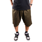 A person stands with hands in pockets, showing only their lower body, dressed in a black T-shirt, Texas Icon Brown Southside 94 Shorts in brown, black socks, and white and black sneakers with purple accents.