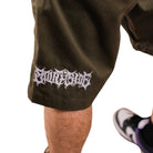 A close-up of a person wearing Texas Icon Brown Southside 94 Shorts with a white embroidered logo on the leg, standing in sneakers featuring black, white, and purple details.