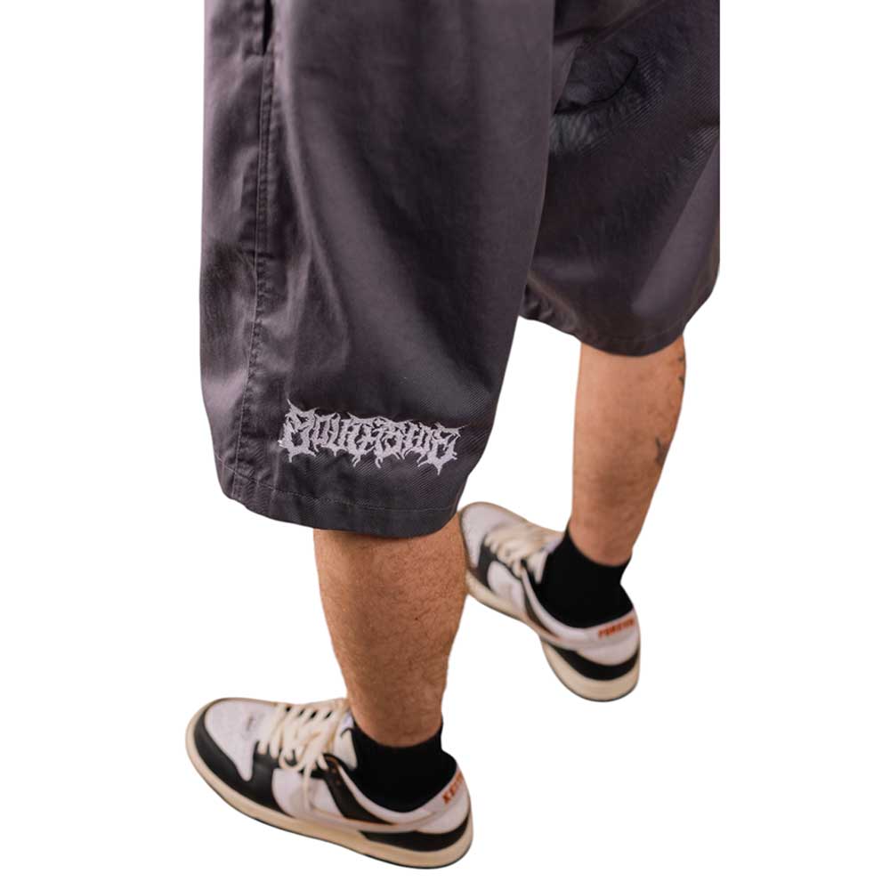 Someone wearing loose, dark gray Texas Icon Gray Southside 94 Shorts, featuring white gothic text on one leg—ideal for skateboarding—paired with black socks and white sneakers with black and orange accents, stands against a white background.