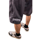 Someone wearing loose, dark gray Texas Icon Gray Southside 94 Shorts, featuring white gothic text on one leg—ideal for skateboarding—paired with black socks and white sneakers with black and orange accents, stands against a white background.