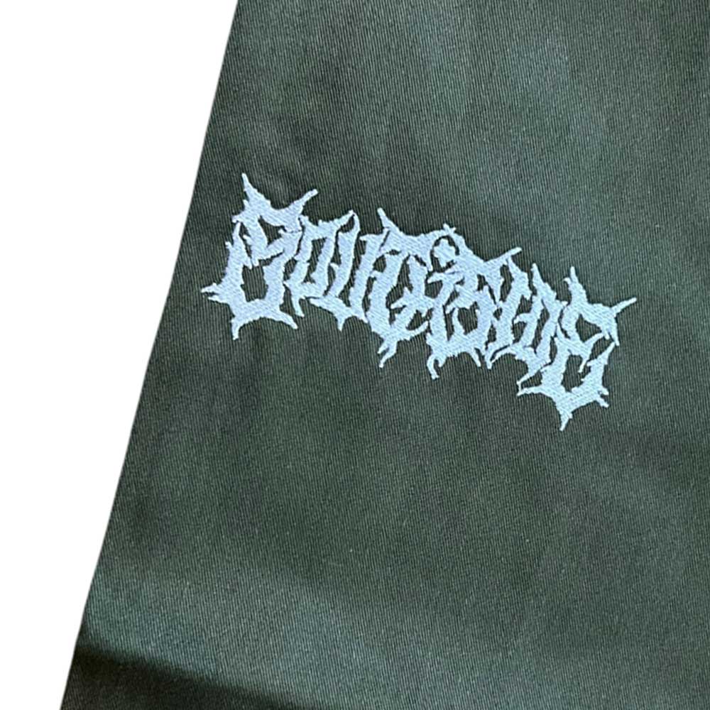 A close-up of dark fabric from the Texas Icon Dark Green Southside 94 Pants, featuring the word OUTCAST embroidered in jagged, white, gothic-style lettering on a plain background.