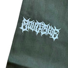 A close-up of dark fabric from the Texas Icon Dark Green Southside 94 Pants, featuring the word OUTCAST embroidered in jagged, white, gothic-style lettering on a plain background.