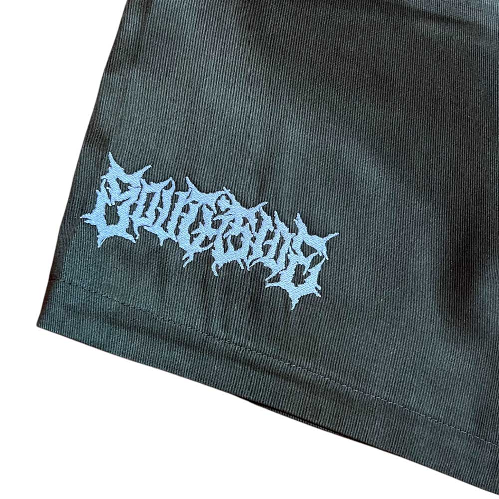 A close-up of dark olive fabric from the Texas Icon Dark Olive Southside 94 Shorts, showing a stylized spiky light blue logo near the hem—evoking the bold look popular in skateboarding circles.