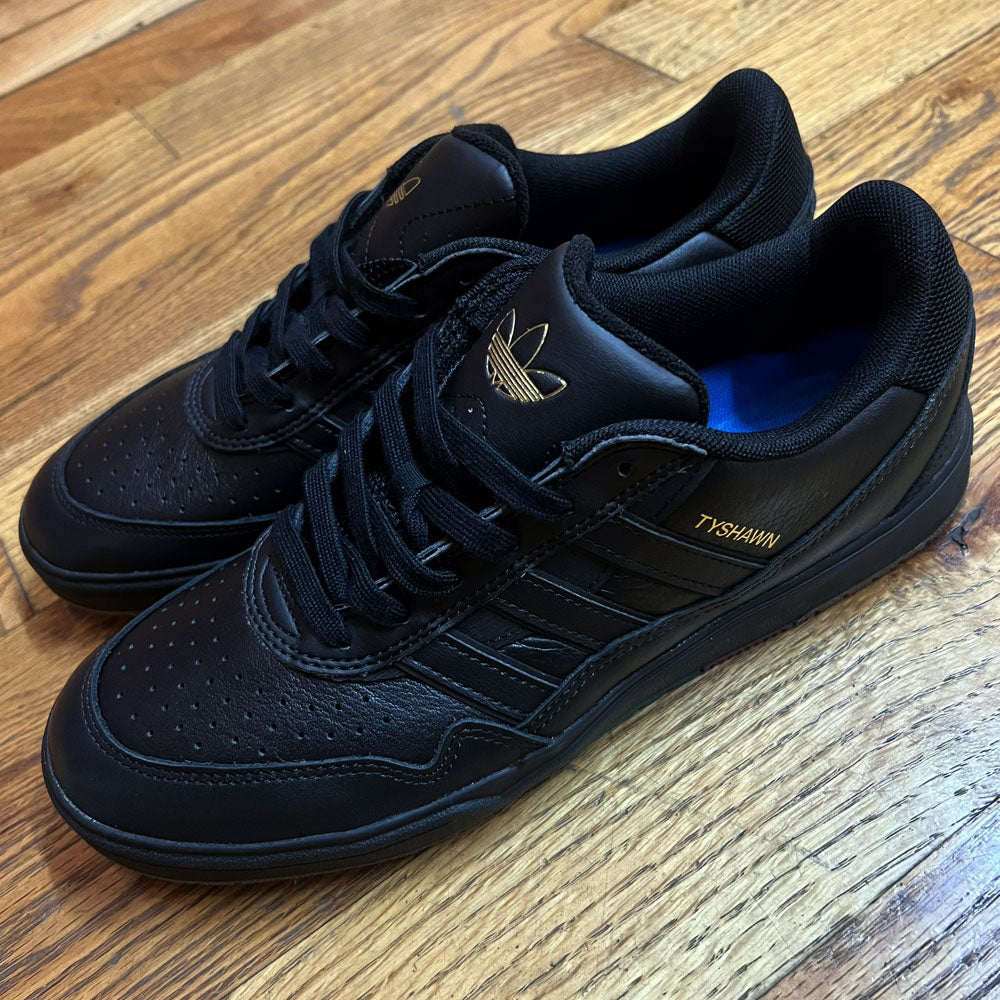 A pair of Tyshawn 2 II Adidas Skateboarding Shoes in Core Black/Core Green leather, with gold branding, black laces, perforated toes, and Adidas logos on tongue and side sits on a wooden floor.