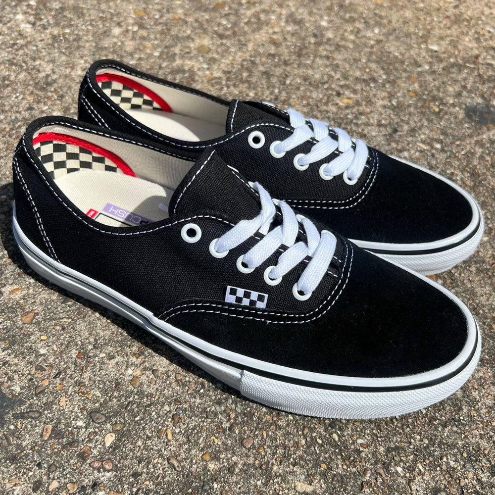 A pair of VNOA5FC8Y28 Suede Canvas Black White Authentic Vans Skateboarding Shoes with white laces and soles rests on textured concrete, featuring a black and white checkerboard detail by the laces and checkered inner lining.