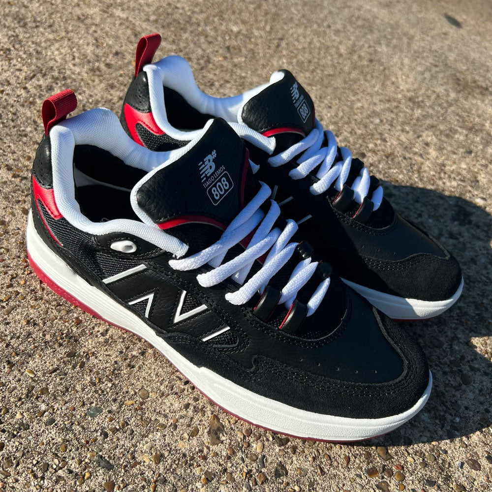 A pair of New Balance NM 808 Tiago Black Red White sneakers with white laces, white midsoles, and red accents sits outdoors on a textured concrete surface in sunlight.