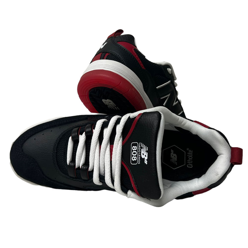 A pair of New Balance NM 808 Tiago Black Red White sneakers with white laces—one upright, one sideways—showcasing Tiago’s signature sole and logo details.