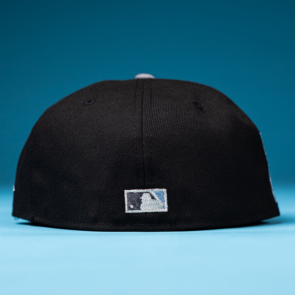 A black The Magician Houston Astros New Era Hat 5950 Fitted is placed on a blue surface, viewed from the back, displaying a small rectangular white and blue logo near the bottom edge.