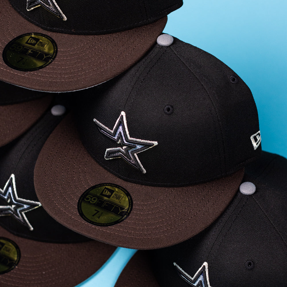 Multiple “The Magician Houston Astros New Era Hat 5950 Fitted” caps in black and brown, each adorned with silver star logos inspired by the Houston Astros, are arranged diagonally on a light blue background. The hats display gold 59FIFTY New Era size stickers on their brims.