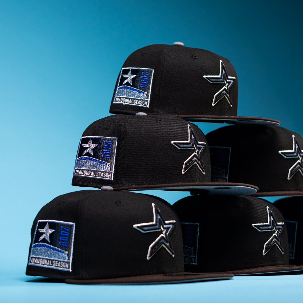 Five black “The Magician Houston Astros New Era Hat 5950 Fitted” caps, featuring a silver star emblem and a blue 2001 Inaugural Season patch, are neatly stacked against a blue background. Offered by Whatafitted and crafted by New Era, these stylish hats blend sleek design with iconic team pride.