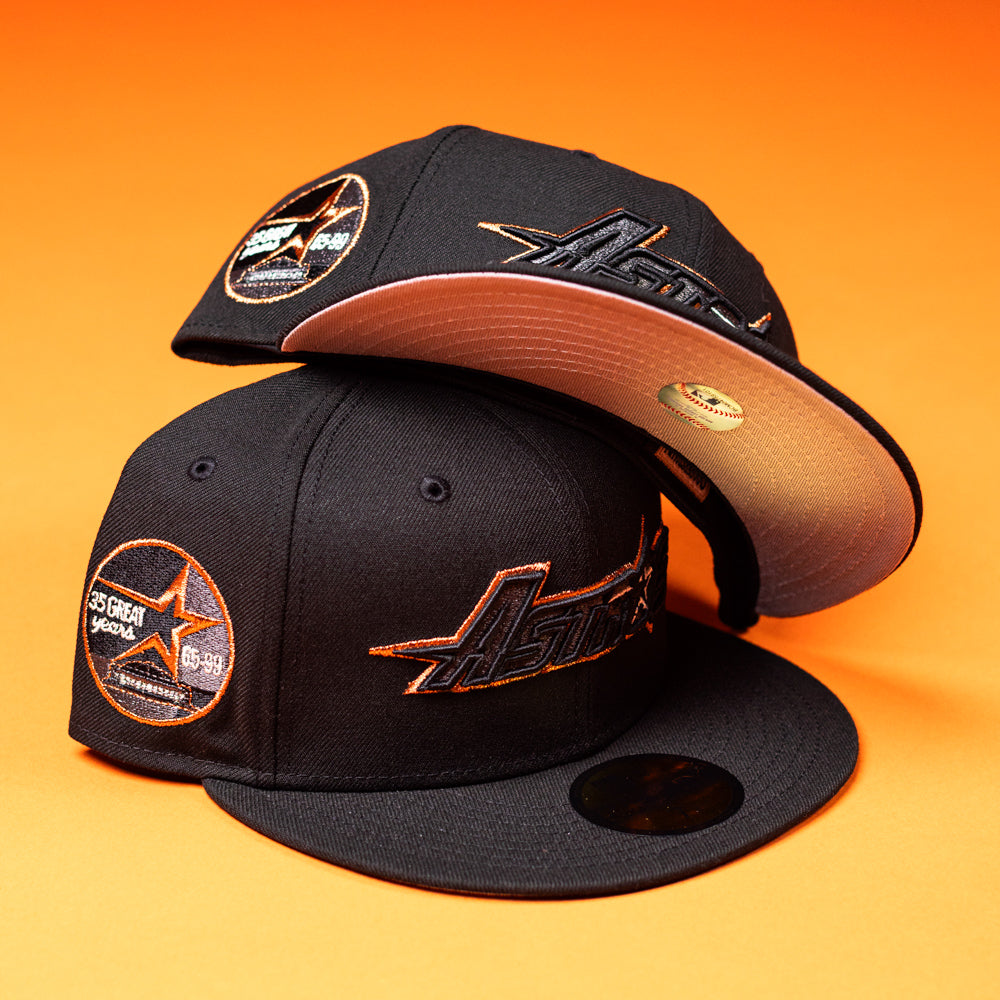 Two BLVCK STAR Houston Astros New Era Hat 5950 Fitted hats in black copper with orange stitching and Astros logo details are stacked on an orange surface, one lying flat and the other resting on top with its brim facing forward.