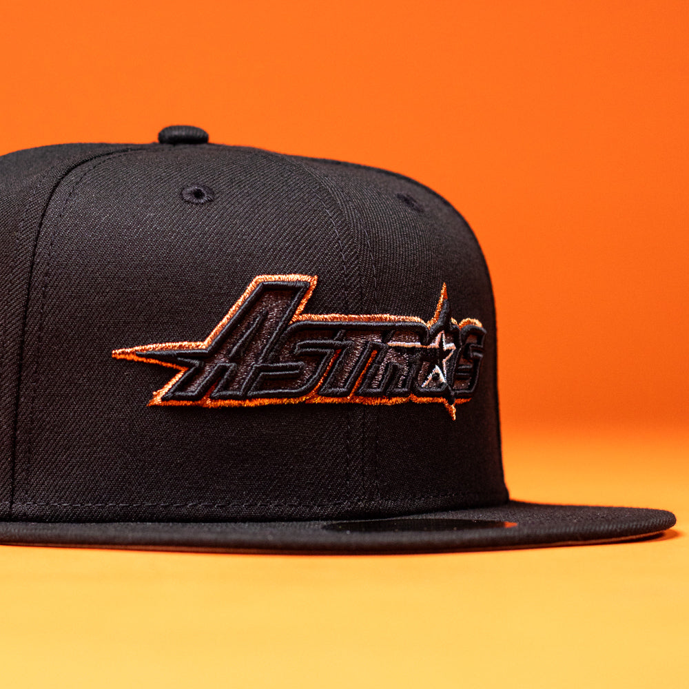 A BLVCK STAR Houston Astros New Era Hat 5950 Fitted in black, featuring “Houston Astros” embroidered in bold black and copper lettering on the front, shown against an orange background.