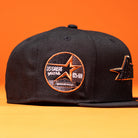 A BLVCK STAR Houston Astros New Era Hat 5950 Fitted in black features an orange and white embroidered patch that reads “35 Great Years 65-99” alongside a star logo inspired by the Houston Astros. Showcasing subtle Black Copper accents, the cap is displayed against an orange background.