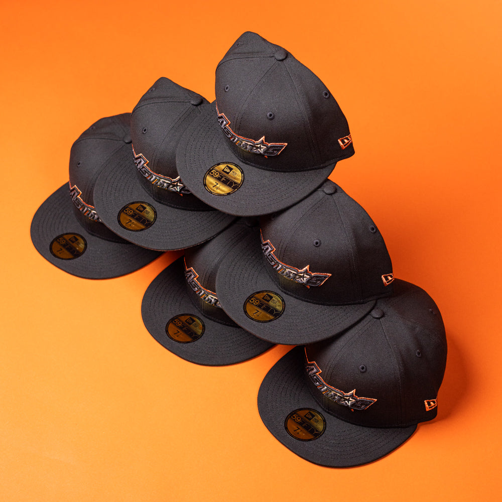 Six BLVCK STAR Houston Astros New Era 5950 Fitted hats in black, each featuring a colorful Astros logo, are stacked in a pyramid shape on an orange background. These hats have gold stickers on their brims.