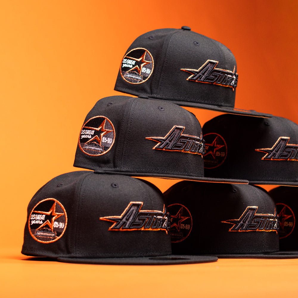 Six BLVCK STAR Houston Astros New Era 5950 Fitted hats are stacked in a pyramid formation against an orange background. These black caps, designed by New Era, showcase striking copper-orange accents along with the iconic team logo and a commemorative patch on the side, giving this collection a bold and stylish appearance.