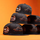 Six BLVCK STAR Houston Astros New Era 5950 Fitted hats are stacked in a pyramid formation against an orange background. These black caps, designed by New Era, showcase striking copper-orange accents along with the iconic team logo and a commemorative patch on the side, giving this collection a bold and stylish appearance.
