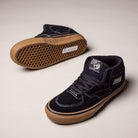 A pair of Vans Half Cab Black Gum White VN0A5FCDB9K suede skate shoes: one upright to show the side and top, the other flipped to display the tan rubber sole. Features include white stitching and visible “Half Cab” branding.