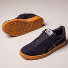 A pair of Japan Pro Asics Black Tan Presido 1201A920003 suede skateboarding shoes with brown gum soles are displayed on a light surface; one stands upright while the other lies on its side, revealing the sole and tread pattern.