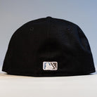 The Eagle Claw Canvas Black Corpus Christi Hooks New Era Hat 5950 Fitted is pictured from the back, displaying the MLB logo in white, blue, and red stitching near the bottom edge against a softly lit plain background.