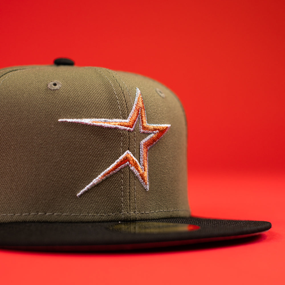 A close-up of The Original Olive Black CC Astros New Era Hat 5950 Fitted, featuring an embroidered orange and white star on the front, a black brim, and set against a vibrant red background.
