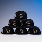 Six Eagle Claw Canvas Black Corpus Christi Hooks New Era Hat 5950 Fitted caps are stacked in a pyramid. Each features a silver embroidered front logo and a colorful side patch. The background transitions from gray to blue.