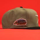 The Original Olive Black CC Astros New Era Hat 5950 Fitted rests on a red surface, featuring an “AUTODROME DRUMMONDVILLE” orange and black patch on the side. The background is solid red.