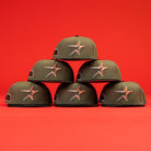 Six The Original Olive Black CC Astros New Era Hat 5950 Fitted caps, featuring the orange Astros star logo, are stacked in a pyramid against a bright red backdrop.