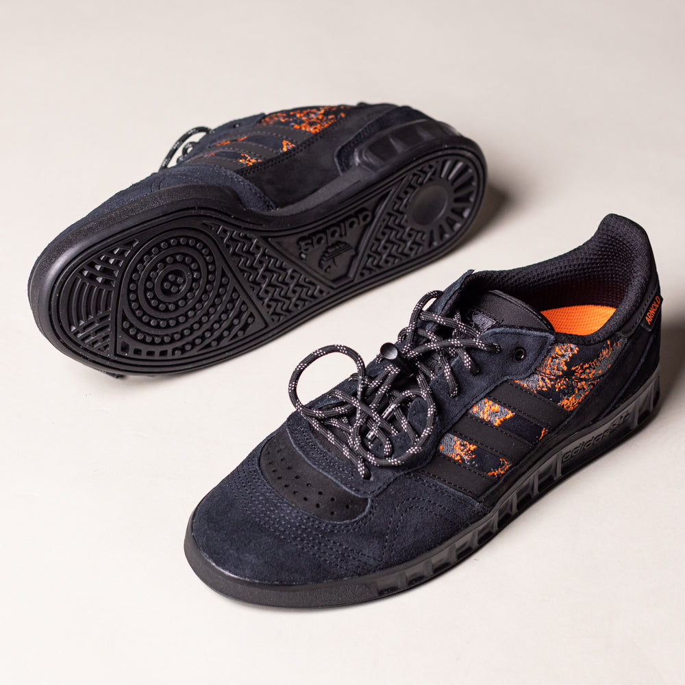 A pair of Handball Top x Mike Arnold Shoes in core black suede with orange details and a textured sole, one upright and one on its side showing the sole, displayed on a light surface—highlighting adidas skateboarding style.