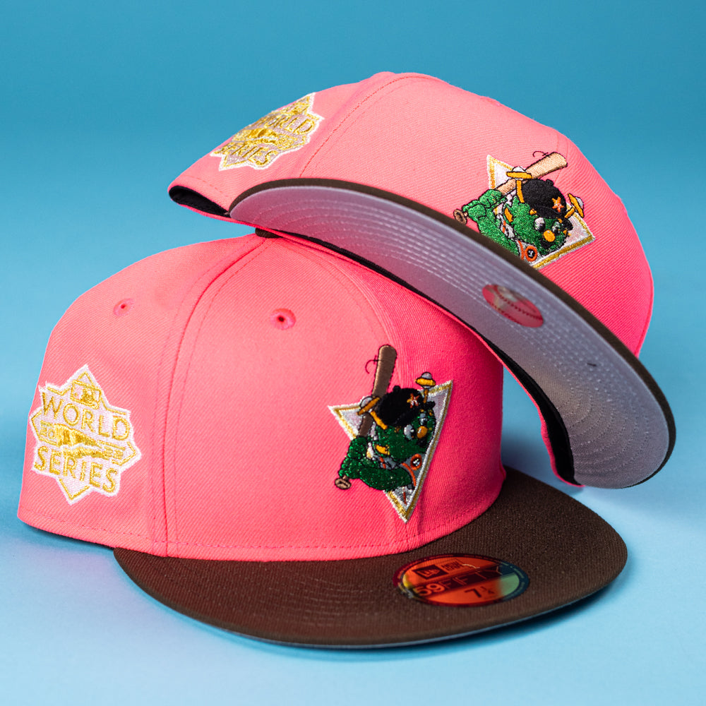 Two Pink Comet WS22 Houston Astros New Era Hat 5950 Fitted caps with brown brims are stacked on a blue surface, each featuring a World Series patch and a green mascot swinging a bat embroidered on the side.