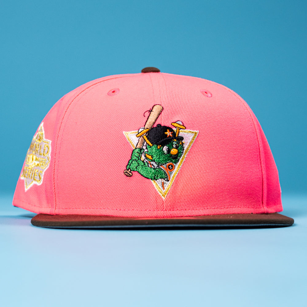 The Pink Comet WS22 Houston Astros New Era Hat 5950 Fitted features a pink crown, black brim, and an embroidered green monster with a bat and cap on the front, all set against a blue background.