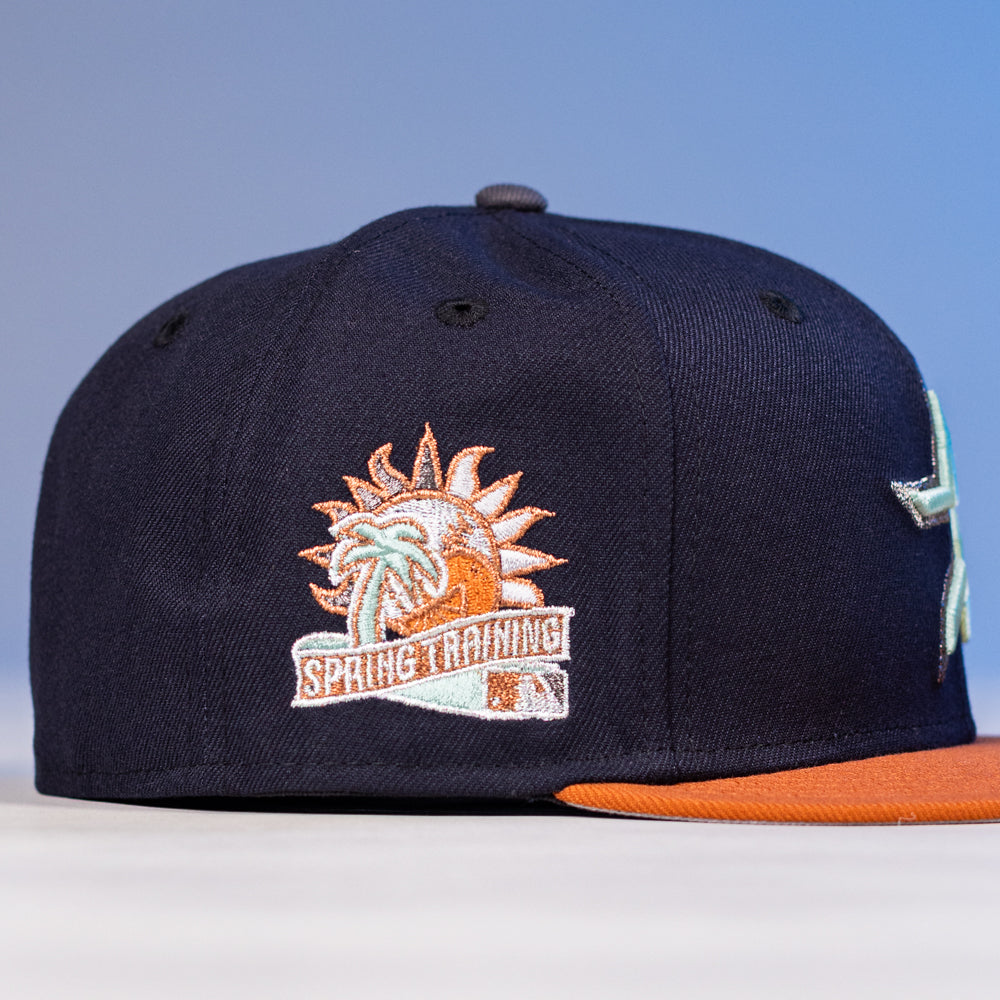 The Feast Navy Rust Houston Astros New Era Hat 5950 Fitted features a navy crown, rust brim, embroidered sun and palm tree logo, and "Houston Astros Spring Training" stitching on the side.