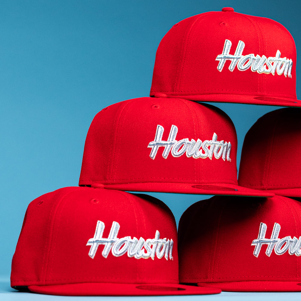 A stack of Cougar Scarlet Houston Cougars New Era Hat 5950 Fitted hats, bright red with "Houston" in white script, is arranged against a solid light blue background.