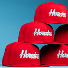 A stack of Cougar Scarlet Houston Cougars New Era Hat 5950 Fitted hats, bright red with "Houston" in white script, is arranged against a solid light blue background.