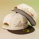 Two Oompa Pink Box Houston Astros 5950 Fitted New Era Hats are stylishly displayed, featuring cream color, brown brims, embroidered Astros logos, and a colorful side patch—one cap upright and the other leaning on top.